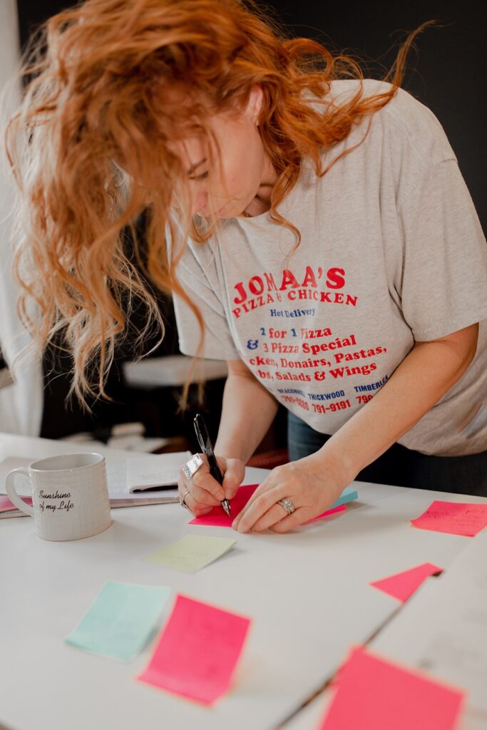 Candace Rae working diligently at her desk, surrounded by colorful sticky notes filled with lists and reminders. She's wearing a casual shirt from her husband's favorite pizza joint in Fort Mac, adding a personal touch to her workspace.